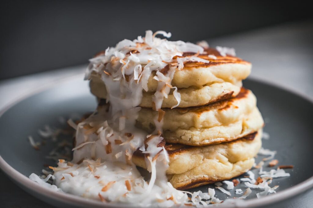Triple stack of a copycat Koko Head Cafe Coconut Ricotta Pancakes drizzled with coconut syrup and toasted coconut shreds